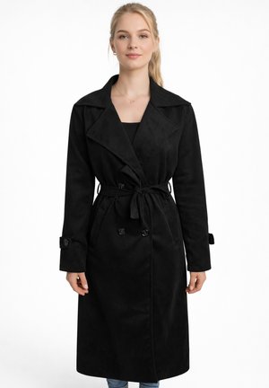 Jonge vrouw met blond haar in een paardenstaart, gekleed in een lange zwarte trenchcoat met ceintuur over een zwart topje, staand tegen een witte achtergrond.