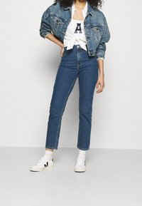 Denim jacket with a relaxed fit, light blue wash; white t-shirt with black letter 'A'; high-waisted blue jeans; white sneakers with black accents.