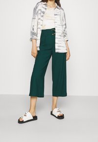 Gröna vida byxor med bälte i midjan, kombinerat med en tie-dye skjorta och vita sandaler med sula i svart och metalliska detaljer.