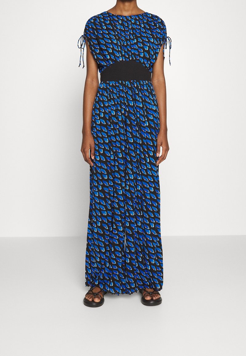 Femme portant une longue robe bleue à motifs avec une ceinture noire à la taille et des manches courtes nouées aux épaules, associée à des sandales noires.