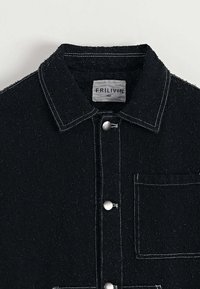 Veste noire en tissu texturé avec un devant à boutons, un col pointu et une poche poitrine. Présente des coutures blanches contrastées.