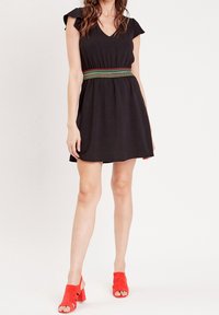 Schwarzes V-Ausschnitt-Kleid mit kurzen Rüschensäumen und einem elastischen, gewebten Gürtel mit roten und grünen Akzenten. Kombiniert mit roten, wadenhohen Sandalen.