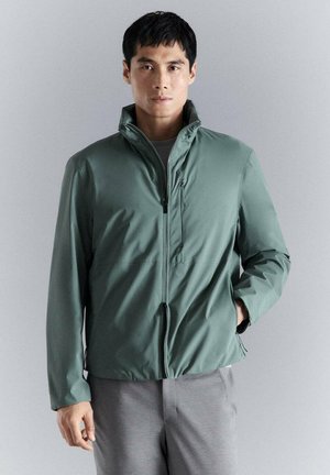 Hombre con chaqueta verde con cremallera y cuello alto, de pie contra un fondo gris neutro, con una mano en el bolsillo.