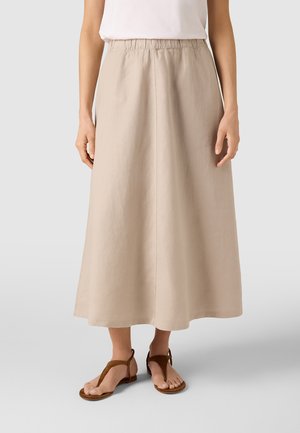 Kvinde iført en beige midi-nederdel med elastik i taljen og brune flade sandaler med ankelremme, stående mod en ensfarvet lys baggrund.