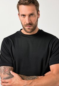 Černé tričko s krátkým rukávem, s texturovaným povrchem, kulatým výstřihem a volným střihem. Model má na pažích tetování a paže má zkřížené.