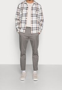Camicia flanella a quadri in beige, blu e bianco, con due tasche frontali; abbinata a pantaloni grigi e sneakers beige chiaro.