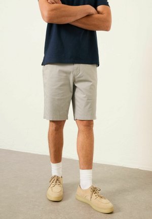 Ein Mann steht mit verschränkten Armen, trägt beige Shorts, ein marineblaues Hemd, weiße Socken und beige Freizeit-Schuhe auf einem grauen Boden.