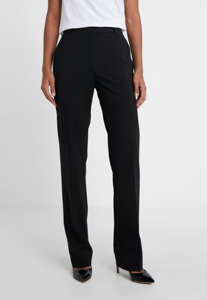 Femme portant un pantalon droit noir, une chemise blanche et des talons hauts noirs pointus, debout devant un fond clair uni.