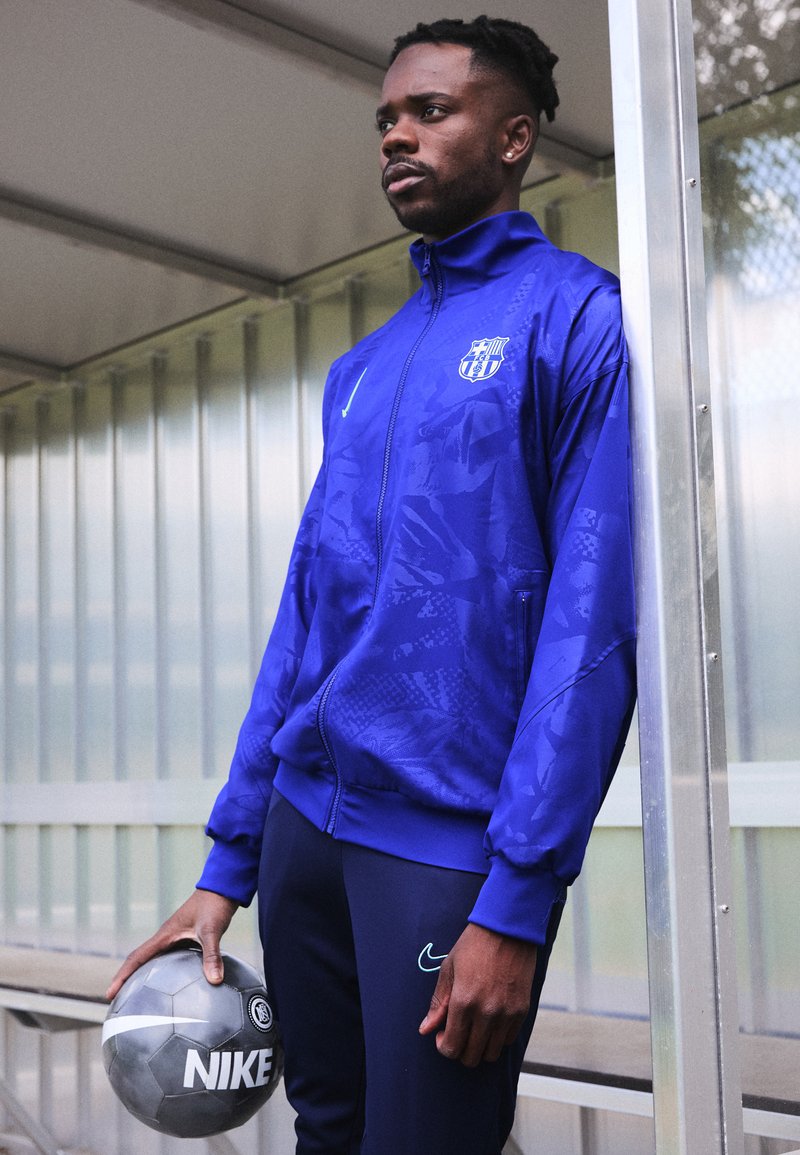 Blauwe sportjack met textuurpatronen en een logo, gecombineerd met navy broeken; houdt een zilveren Nike voetbalschoen vast.