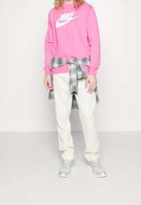 Rosa Nike-tröja med vit logga, grå rutig flanell skjorta knuten i midjan, krämfärgade byxor och grå sneakers. Avslappnad outfit.