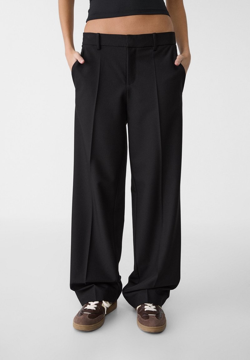 Femme portant un pantalon noir ample avec des plis à l'avant et les mains dans les poches, associée à des baskets marron avec des lacets blancs.