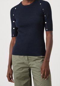 Námořnicky modrý pletený top s krátkými rukávy, zdobený stříbrnými cvočky podél ramen a přiléhavým střihem. Texturovaný žebrovaný materiál.