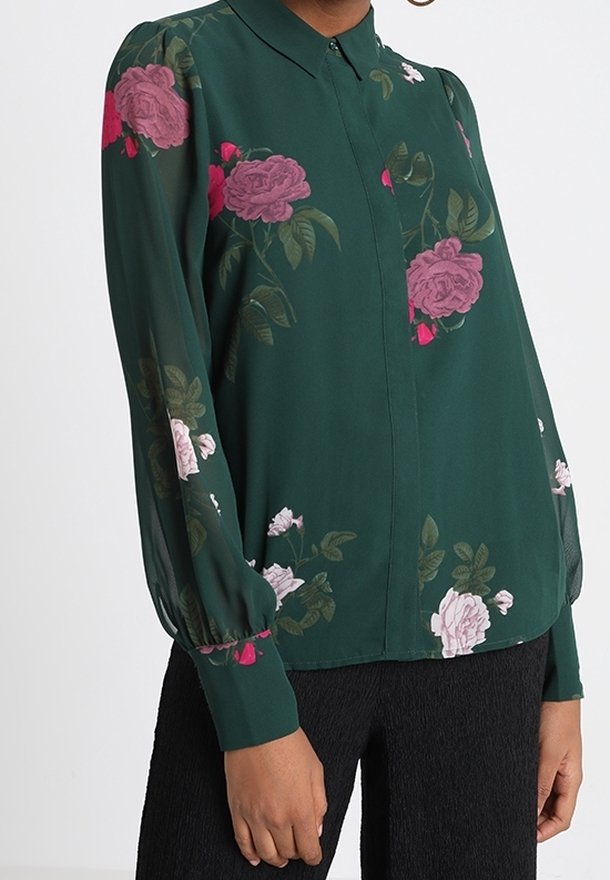 Blouse à manches longues vert foncé avec des imprimés floraux roses et blancs, avec un devant boutonné et des manches bouffantes, portée avec un pantalon noir texturé.