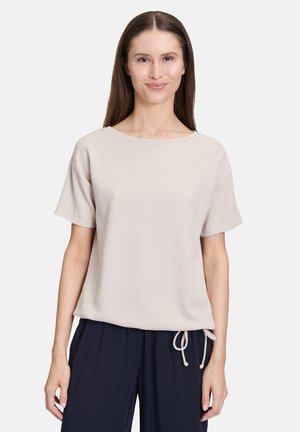 Korte mouwen beige top met een textuurpatroon, relaxte pasvorm, brede halslijn en verstelbare koord aan de onderkant. Gecombineerd met een marineblauwe broek.