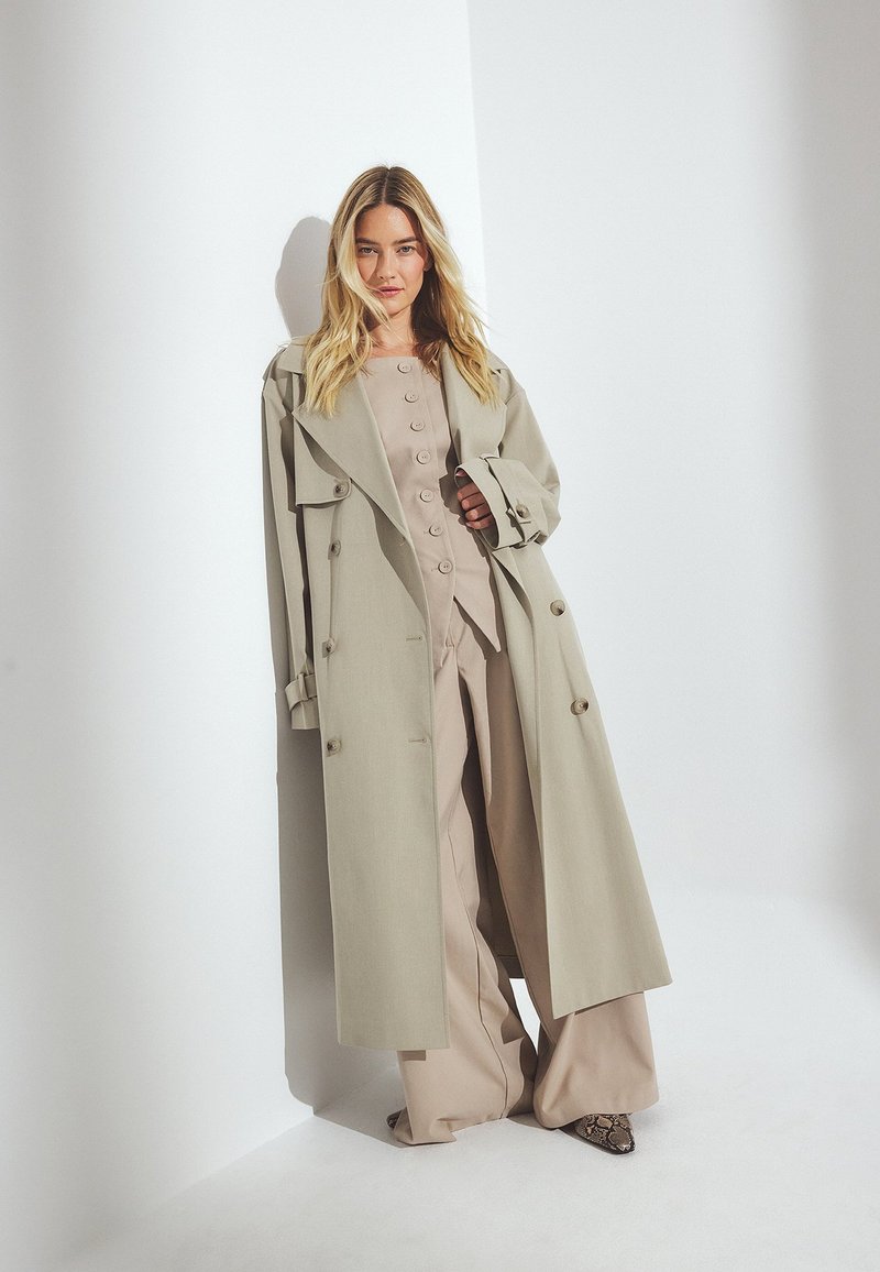 Bežs lietussargs ar divām pogu rindām, plašiem atloka galiem un pogu aizdari, uzvilkts virs atbilstoša bežīga tērpa. Teksturēti čūskas raksta apavi.