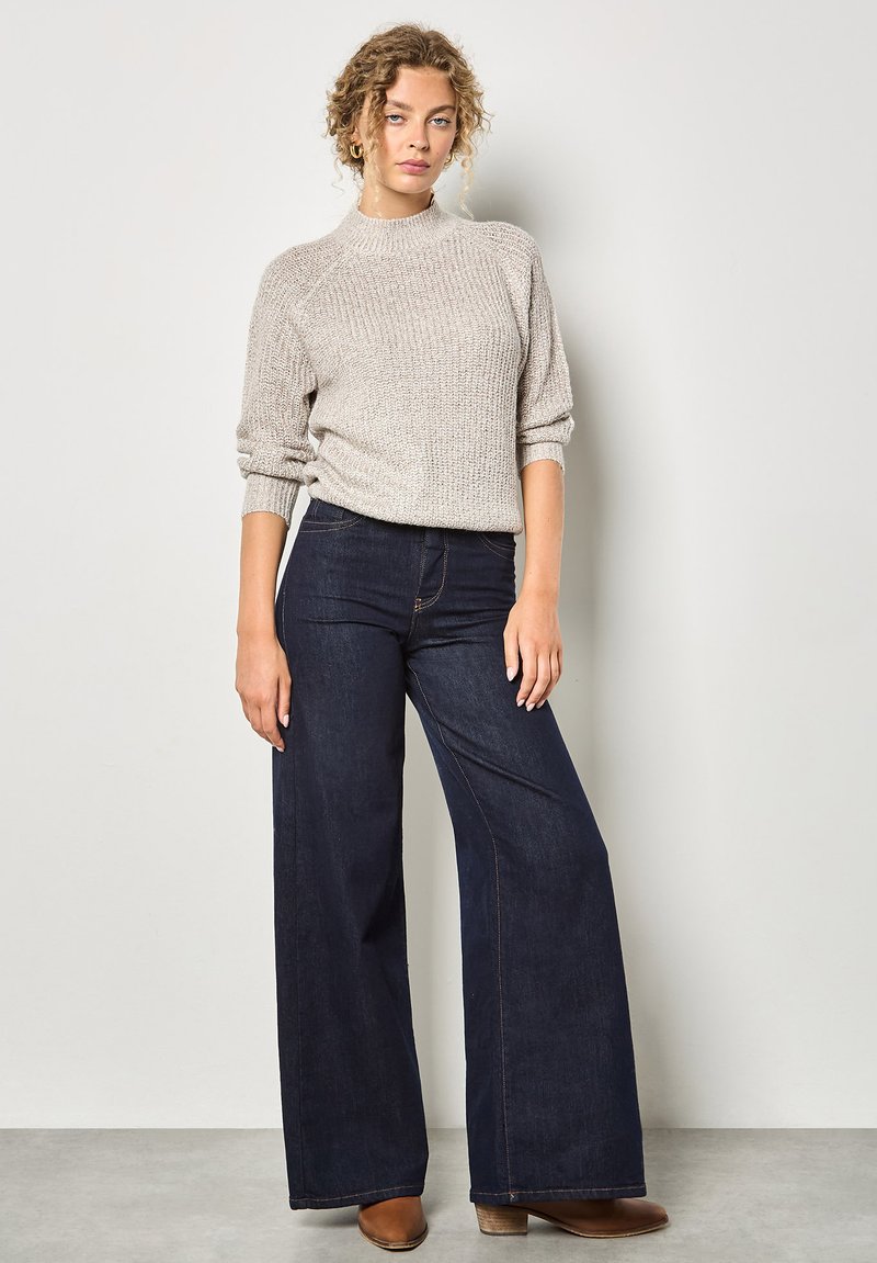 Hellgrauer Strickpullover mit Ripptextur, kombiniert mit weiten dunkelblauen Jeans mit kontrastierenden Nähten und einfarbigen braunen Stiefeletten.