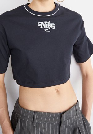 Osoba nosi czarny krótki t-shirt Nike z białym logo i szare paskowane spodnie z wysokim stanem, pokazując brzuch i pępek.