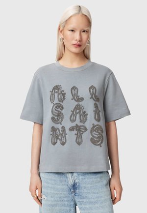Jeune femme aux longs cheveux blond platine portant un t-shirt gris surdimensionné avec des lettres décoratives noires et un jean bleu clair.