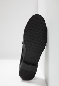 Svart lackskofunktion med texturerat grepp, som har ett upprepat logomönster, platt design och slät yta. Ljus reflekteras från ytan.