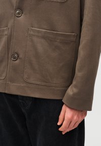 Brown canvas jacket featuring a button closure, two large patch pockets, and straight sleeves, paired with dark denim pants.