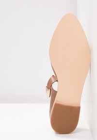 Ruskea nahkakenkä, jossa on terävä kärki, kultainen yksityiskohta ja sileä, beige pohja, jossa on tekstuuri.