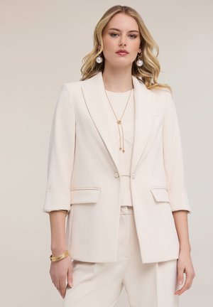 Blondýnka v béžovém saku a kalhotách na míru, s dlouhými perlovými náušnicemi, zlatým náhrdelníkem a několika zlatými náramky na levém zápěstí.