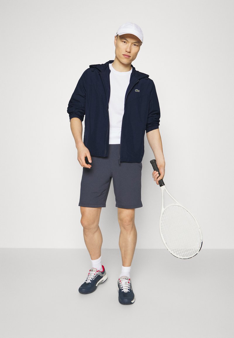 Marinefarbener Zip-Up-Jacke, weißes T-Shirt, graue Shorts und blaue Tennisschuhe mit pinken Akzenten; hält einen Tennisschläger. Minimalistisches Design.
