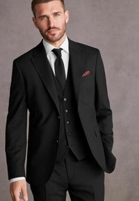 Costum negru în trei piese, cu un croi slim, având rever cu tăietură, vestă cu șase nasturi și o batistă roșie, completat cu o cămașă albă și o cravată.