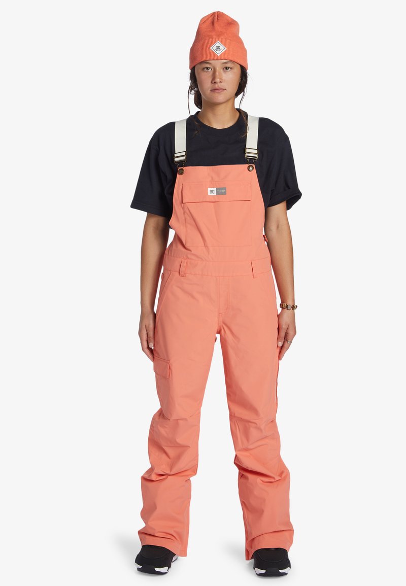 Oranje sneeuwoverall met verstelbare schouderbanden, zijzakken en een taps toelopend ontwerp, gecombineerd met een zwart T-shirt en een oranje muts.