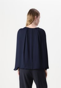 Blusa fluida blu navy con scollo rotondo e maniche larghe in stile cappotto. Tessuto liscio con dettagli plissettati sul retro.
