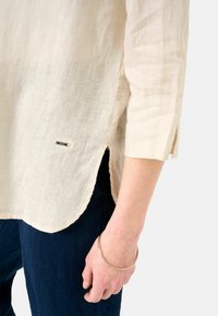 Jasno beżowa lniana koszula z rękawami o długości trzech czwartych, z bocznymi rozcięciami u dołu oraz małym srebrnym detalem w lewym dolnym rogu.