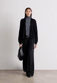 Črna pletena jakna preko sive turtleneck majice, v kombinaciji s širokimi črnimi hlačami in črno torbico. Čist, minimalističen dizajn.
