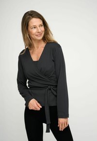 Fekete szövött felső hosszú ujjal, V-kivágással és állítható megkötővel a deréknál. Puha anyag és sima textúra. Fekete tanktopra van rétegezve.