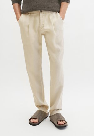 Person trägt beige plissierte Hose und braune offene Pantoletten, steht mit den Händen in den Taschen vor weißem Hintergrund.