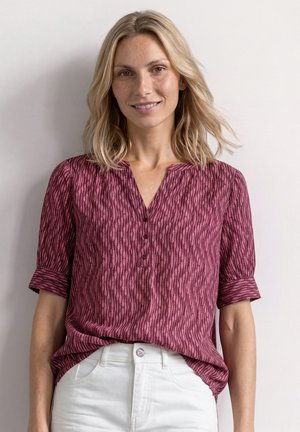 Femme blonde portant un chemisier rouge à manches courtes à motifs avec des boutons et un pantalon blanc, debout devant un fond clair uni.