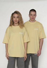 Zwei Models tragen übergroße gelbe T-Shirts mit kurzen Ärmeln. Auf jedem Shirt steht in Weiß auf der Brust das Wort „MULTIPLY“.