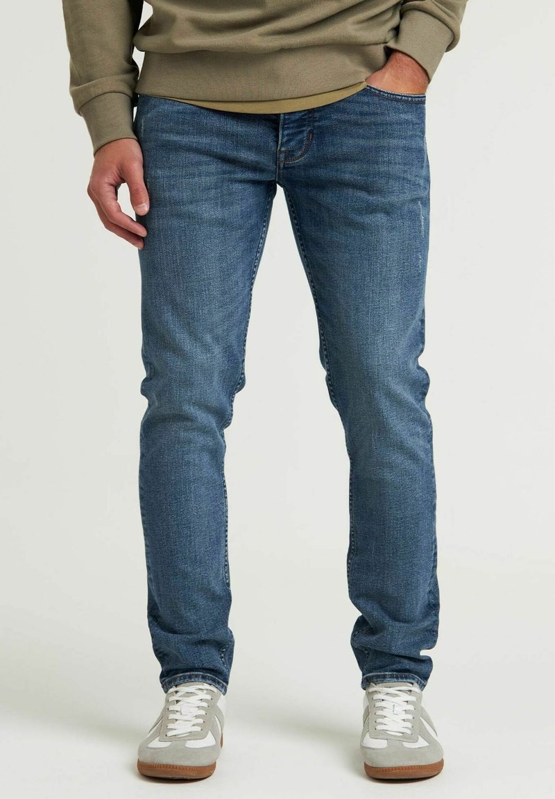 Homme portant un jean bleu slim, un sweat beige et des baskets blanches, debout avec une main dans la poche devant un fond uni.