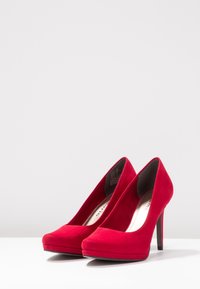 Červené semišové vysoké podpatky s špičatou špičkou, platformou vpředu a tenkým jehlovým podpatkem. Hladký povrch a minimalistický design bez viditelných prvků.