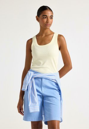 Kvinde iført cremefarvet tanktop og lyseblå shorts med en stribet skjorte bundet om livet, stående mod en ensfarvet hvid baggrund.