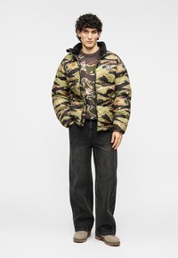 Επενδεδυμένο καμουφλάζ μπουφάν σε πράσινο, καφέ και μαύρο χρώμα. Συνδυασμένο με πουλόβερ με σχέδιο, μαύρα τζιν με φαρδιά πόδια και μπεζ παπούτσια.