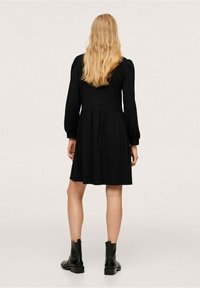 Robe noire, manches longues bouffantes, corsage ajusté, jupe évasée, texture douce. Portée avec des bottines noires à la cheville, design minimaliste.