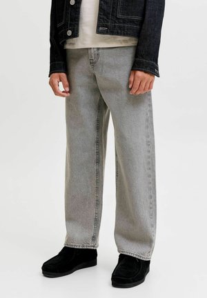 Person, die hellgraue Jeans mit geradem Bein, schwarze Schuhe, ein weißes Hemd und eine dunkle Jeansjacke trägt, steht vor einem einfarbigen Hintergrund.