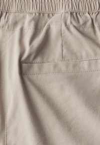Pantaloni corti in tessuto beige, con una tasca posteriore e vita elastica. La texture è liscia con una leggera lucentezza. I dettagli delle cuciture sono visibili.