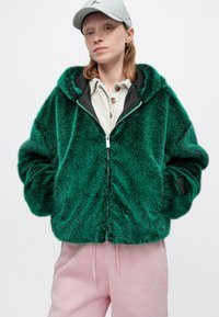 Grüne Kunstfelljacke mit Reißverschluss, Kapuze und seitlichen Taschen. Getragen über einem hellen Hemd mit Kragen und rosafarbenen Jogginghosen.