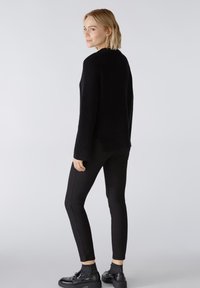 Schwarzer gerippter Pullover mit langen Ärmeln, kombiniert mit schwarzen enganliegenden Jeans und Schnürschuhen. Der Pullover hat einen hohen Kragen und seitliche Schlitze.