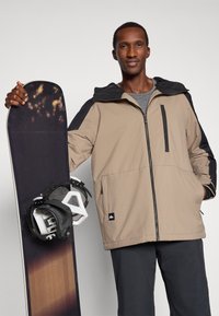 Άνδρας με μπεζ και μαύρο μπουφάν και σκούρες παντελόνες κρατάει ένα σνόουμπορντ σε όρθια θέση με το ένα χέρι, με φόντο απλό.