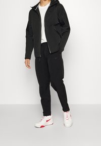 Chaqueta negra con cremallera y capucha, pantalones negros y zapatillas blancas con detalles rojos. El material parece ligero, con logotipos de Nike en la ropa.