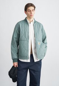 Πράσινο bomber μπουφάν με ραμμένη επένδυση, διαθέτει πλεκτό γιακά, δύο εμπρόσθιες τσέπες και κλείσιμο με φερμουάρ, συνδυασμένο με λευκό πουκάμισο και σκούρο παντελόνι.