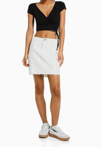 Černý zúžený crop top, bílá džínová sukně s roztrhaným lemem a kapsami ve stylu cargo. Modelka má na sobě bílé tenisky s černými a béžovými akcenty.