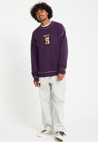 Paarse sweatshirt met een centraal grafisch label, met een beige illustratie. Gecombineerd met lichte denim jeans en neutrale sneakers.